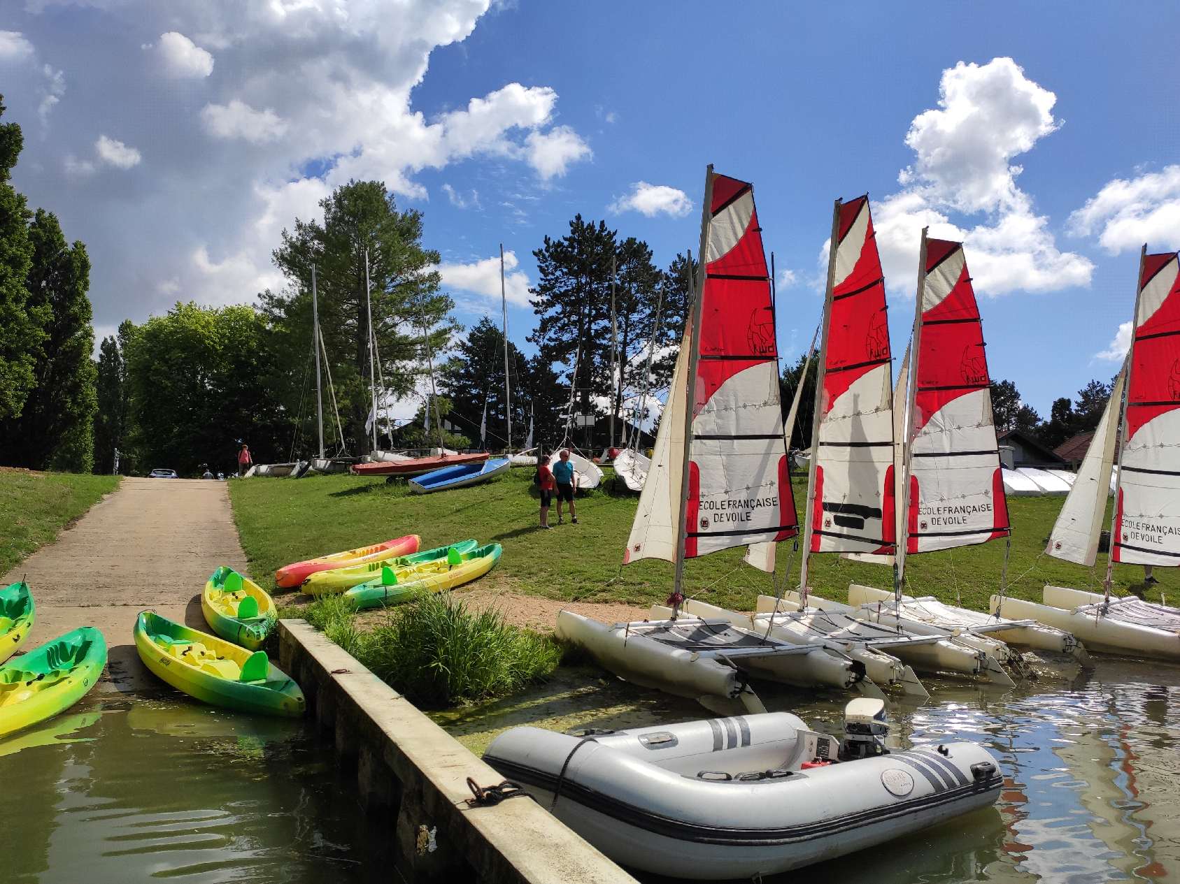 Base nautique : voile légère et paddle au bord de l'eau Catamarans de sport à voiles rouges et blanches alignés sur un ponton, avec kayaks et paddles gonflables au bord d'un plan d'eau entouré d'arbres