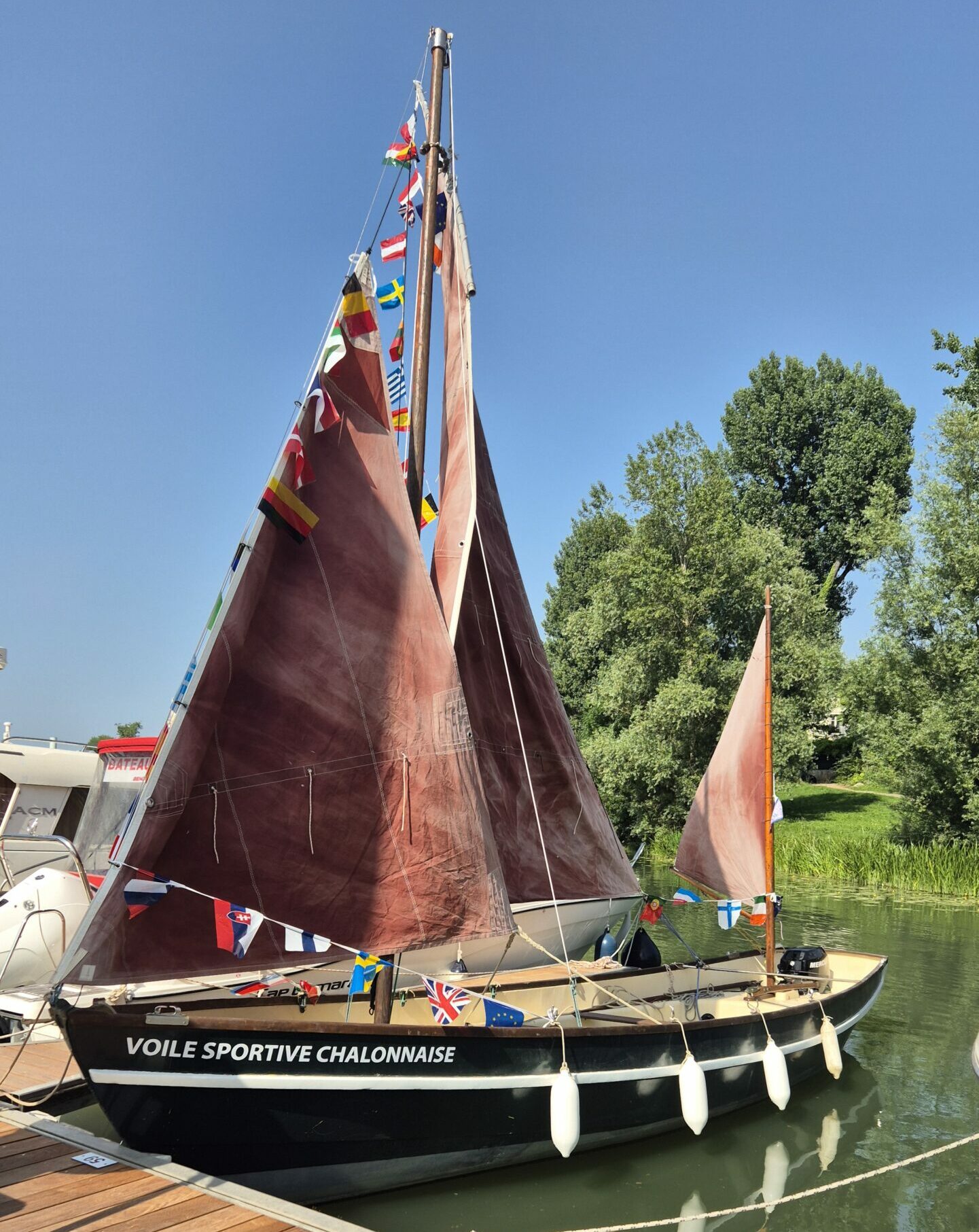 Balade en vieux gréement Voilier en bois à deux voiles brunes naviguant sur une rivière calme avec des passagers à bord, entouré d'une rive boisée